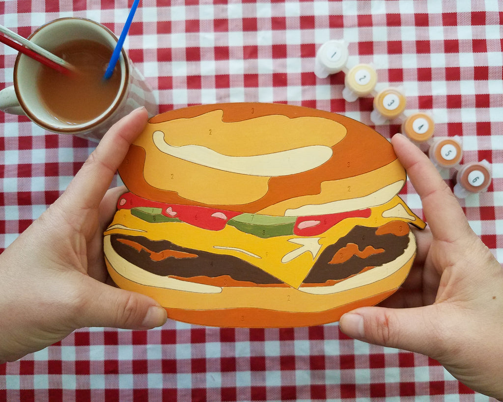 The Cheeseburger Paint by Number Kit features a tasty design that will make your mouth water! A toasty bun sandwiches a burger patty covered in golden melted cheese, a few bright green pickles, covered in vibrant red ketchup. A great craft kit for all ages and aspiring artists.